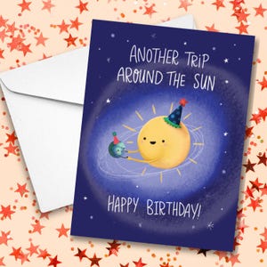 May include: A birthday card with a cartoon sun wearing a party hat, holding a small planet. The card has the text "Another Trip Around the Sun" and "Happy Birthday!" on a dark blue background with stars. A white envelope is included.