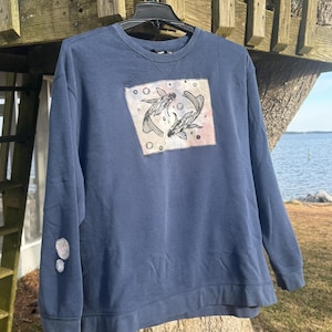 May include: A navy blue sweatshirt with a square graphic featuring two koi fish. The graphic is in shades of black, white, and pink. The sweatshirt is hanging on a black hanger outdoors.