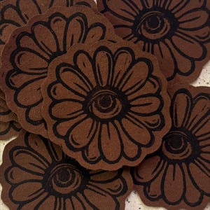 May include: A collection of brown, flower-shaped coasters with a black eye design in the center. The coasters have a textured appearance and are arranged in a slightly overlapping pattern. The floral design features curved petals.