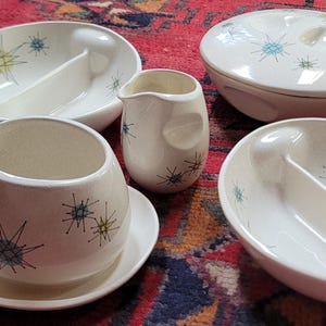 May include: A collection of off-white ceramic dinnerware featuring a starburst pattern in blue and green. The set includes two divided oval bowls, a lidded round casserole dish, and two creamers, one of which is placed on a matching saucer.