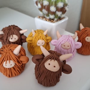 May include: Six handmade miniature Highland Cow figurines, each with a different colour of fur: brown, orange, yellow, pink, brown, and dark brown. The figurines are made of polymer clay and have a fuzzy texture.