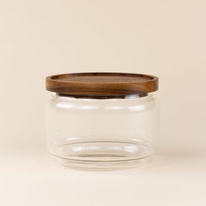 May include: A clear glass jar with a wooden lid. The jar is cylindrical and has a wide mouth. The lid is made of dark wood and has a smooth, rounded top.