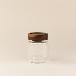 May include: A clear glass storage jar with a cylindrical body and a wide, flat, dark brown wooden lid. The jar features a smooth, transparent glass construction with a slightly tapered base and a thick, polished wooden cap on top.