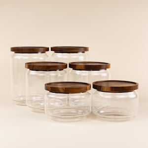 May include: Five glass jars with wooden lids. The jars are clear glass and the lids are a dark brown wood. The jars are arranged in a row from largest to smallest.