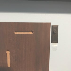 May include: The back of a wooden picture frame with two hanging slots and a black plastic hook attached to the wall.