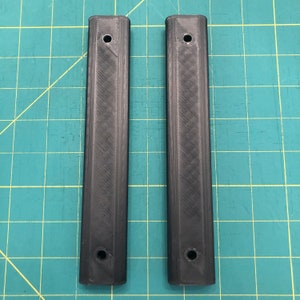 Może przedstawiać: Dwa czarne prostokątne elementy z tworzywa sztucznego z fakturowaną powierzchnią i otworem na każdym końcu.