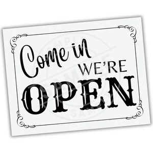 May include: A rectangular white sign with black text that reads "Come in WE'RE OPEN". The sign has decorative flourishes in the corners and a black border. The text is in a mix of cursive and bold fonts.