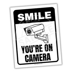 May include: A black and white sign with the text "SMILE YOU'RE ON CAMERA" and a security camera graphic. The sign has a black border and is on a white background. The website address is at the bottom.