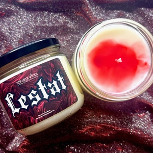 May include: A glass candle jar with a red and white wax candle inside. The jar has a black lid and a label that says "Strange Wicks Candle Co." and "Lestat" in a gothic font.