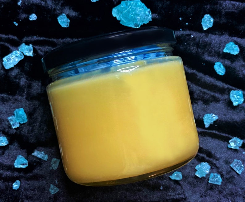 May include: A glass jar with a black lid filled with a yellow substance. The jar is sitting on a black surface with blue crystals scattered around it.