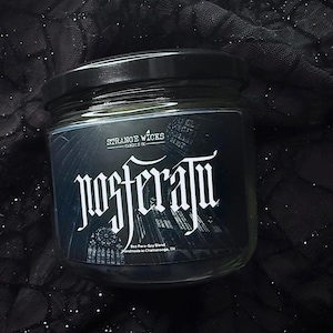 May include: A black glass candle jar with a white label that reads "STRANGE WICKS CANDLE CO." and "NOSFERATU" in a gothic font. The candle is a dark grey color and has a black lid.