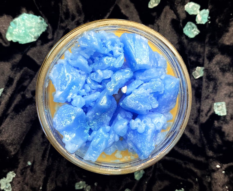May include: A blue candle with a textured, crystal-like surface. The candle is in a clear glass jar with a yellow wax base.