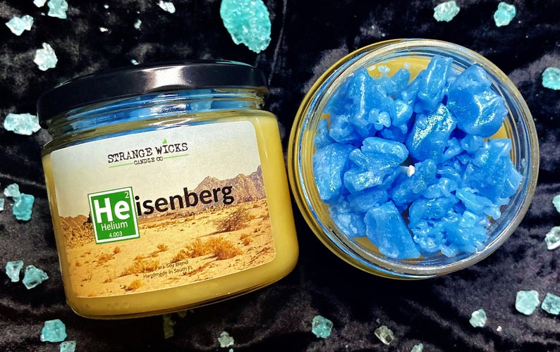 May include: A yellow candle in a glass jar with a label that says "Strange Wicks Candle Co. Heisenberg He Helium 4.003" and a desert landscape. The candle is topped with blue wax crystals.