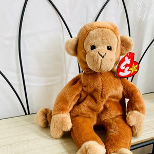May include: A brown plush monkey toy with a tag attached. The monkey has a light brown face, hands, and feet, with a darker brown body. It is sitting on a light-colored wooden shelf.