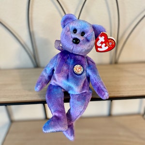 Può includere: Orso di peluche Beanie Baby tie-dye viola e blu con un'etichetta a forma di cuore che dice "Ty Beanie Babies". L'orso indossa un bottone blu con un cuore e le parole "Think of Me" su di esso.