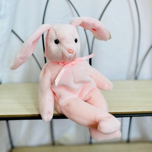 May include: A pink plush bunny rabbit with long ears and a pink ribbon around its neck. The bunny is sitting on a wooden shelf.