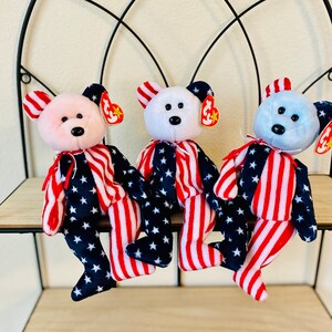 May include: Three plush teddy bears wearing red, white, and blue striped outfits with stars. The bears are sitting on a wooden shelf with a black metal frame.
