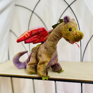Puede incluir: Un peluche de dragón con un cuerpo texturizado en tonos marrones, morados y verdes. Tiene alas rojas iridiscentes, una larga cola morada y una cresta verde. El dragón está posado sobre una estantería de color claro, evocando fantasía.