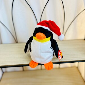May include: A white and black penguin plush toy wearing a red Santa hat with a white pom-pom. The penguin has a yellow beak and orange feet. The toy has a red Ty tag with a white star.