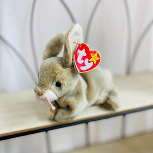 May include: A brown and white plush bunny with a pink nose and a red Ty Beanie Babies tag. The bunny is lying on a wooden shelf.