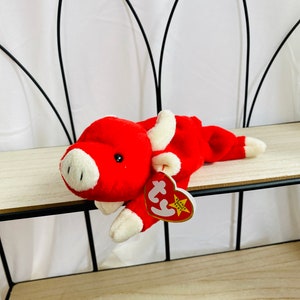 Puede incluir: Juguete de peluche rojo en forma de toro con la cara blanca y la barriga blanca. El juguete tiene una etiqueta Ty unida a su pata. La etiqueta es amarilla con una estrella roja y las palabras "Beanie Baby" impresas en ella.