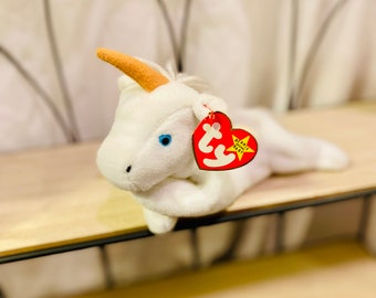 Könnte beinhalten: Weißes Einhorn-Plüschtier mit braunem Horn und blauen Augen. Das Plüschtier liegt auf einem Holzregal mit einem roten "Ty"-Etikett.