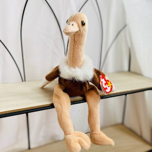 Puede incluir: Un juguete de peluche de avestruz marrón y beige sentado en un estante de madera. El juguete tiene un cuello de piel blanca y una etiqueta Ty roja y blanca que dice "Ty".
