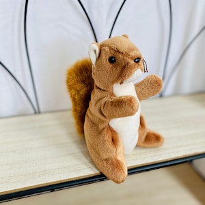 May include: A plush squirrel toy with brown fur, a fluffy tail, and a white belly. The stuffed animal is sitting on a light-colored wooden shelf. The background is a white curtain and a black metal frame.
