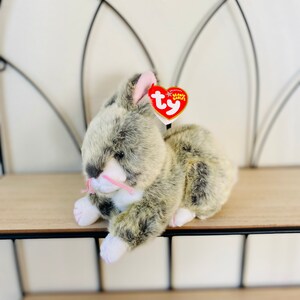 May include: A gray and white plush bunny with pink ears and nose. The bunny is sitting on a wooden shelf with a red Ty Beanie Babies tag attached to its ear. The tag says "Original Beanie Babies".
