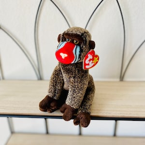 May include: A brown and black stuffed monkey with a red face and blue stripes. The monkey is sitting on a wooden shelf. The monkey has a Ty tag attached to its ear.