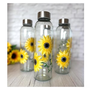 May include: Clear glass water bottles with silver lids, decorated with hand-painted yellow sunflowers. The bottles are cylindrical and stand on a light wooden surface, with sunflowers in the background. A refreshing and decorative item.