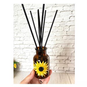 May include: Amber glass diffuser bottle featuring a hand-painted sunflower design. The bottle contains black reeds for fragrance diffusion. The sunflower has yellow petals and a dark centre. The bottle is held in a hand.