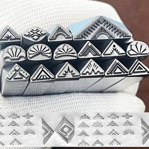 May include: A set of 18 metal stamps with various geometric and floral designs. The stamps are arranged in a stack, with the designs facing up. The stamps are made of a silver-colored metal and have a polished finish.
