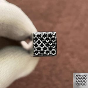 May include: A square metal stamp with a repeating pattern of black and white semi-circles, resembling fish scales. The stamp is held against a blurred background, showcasing the detailed design.