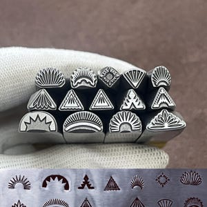 Puede incluir: Un conjunto de sellos metálicos con diversos diseños geométricos y florales. Los sellos se sostienen en una mano enguantada, con una lámina metálica debajo que muestra impresiones estampadas de los diseños. Los diseños incluyen rayos de sol, triángulos y patrones florales estilizados.