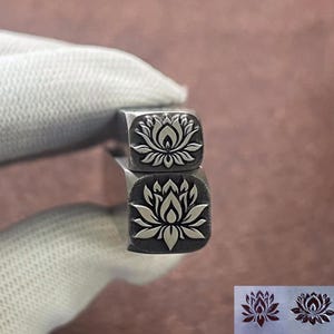 Op de afbeelding: Twee metalen stempels met een lotusbloem ontwerp. De stempels zijn vierkant met een gedetailleerd bloemenpatroon. De stempels worden vastgehouden door een gehandschoende hand. De achtergrond is een vage, neutrale kleur.