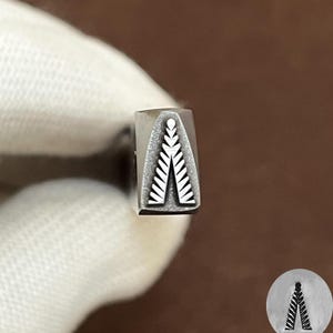 May include: A metal stamping tool with a rectangular shaft featuring a recessed design of a stylized fern leaf or pine branch pattern on the end. The stamp creates a symmetrical, pointed impression with small, repeating leaf-like segments.