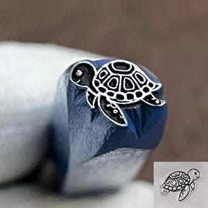 May include: A metal stamping tool featuring a raised turtle design on its end. The turtle has a detailed shell pattern and flippers. A small inset shows two clear impressions of the turtle design stamped onto a flat surface in black ink.
