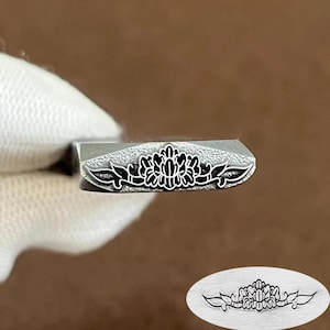 May include: A metal stamping tool featuring a detailed floral engraving on its flat end. The design consists of a central flower with symmetrical leaf-like extensions. An oval inset shows the clear, crisp impression left by the stamp on a surface.