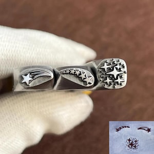 Puede incluir: Una herramienta de estampado de metal con tres diseños distintos: una estrella fugaz, una fila curva de estrellas y un grupo de estrellas. La herramienta se sostiene en una mano enguantada, mostrando los detallados patrones de estrellas. Ideal para manualidades.