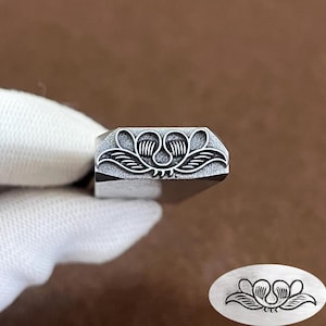 May include: A silver-toned metal stamping tool featuring a recessed floral or leaf-like design on its flat end. The stamp head displays a symmetrical pattern with curved lines and internal striations, accompanied by a separate oval impression.