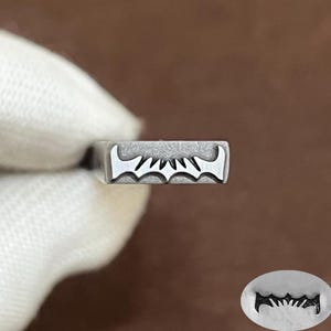 May include: A rectangular metal stamping tool with a brushed silver finish. The face of the tool features a deeply engraved, stylized bat-like emblem with pointed wings and a central peak. An impression of the design is shown in the bottom corner.