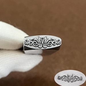 May include: A metal stamping tool featuring a detailed butterfly and scrollwork design on its face. The stamp is shown in profile, and an oval inset displays the clear, embossed impression of the butterfly motif created by the tool.
