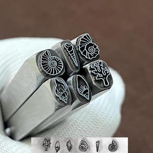 Peut inclure: Un ensemble de six outils de frappe en métal argenté, chacun présentant un motif marin distinct à l'extrémité, incluant divers coquillages, une ammonite en spirale et une branche de corail. Les empreintes des six motifs sont visibles en bas.