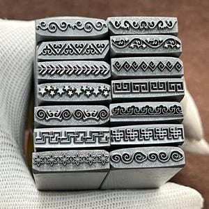 May include: A set of 10 metal stamps with different decorative patterns. The stamps are arranged in a vertical stack and feature designs such as swirls, stars, and geometric shapes.