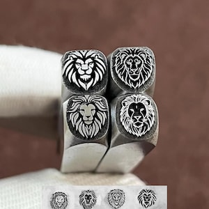 May include: Four metal stamping tools with square heads, each featuring a distinct engraved lion head design. Below the tools, four corresponding impressions of the lion designs are visible on a light surface, showing the detailed results of each stamp.