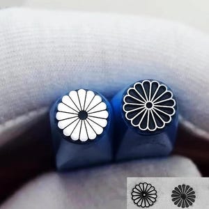 May include: Two blue-handled metal stamps with a chrysanthemum design. One stamp has a white flower on a black background, and the other has a black flower on a white background. The stamps are shown against a soft, white background.