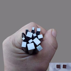 以下が含まれることがあります： 正方形と長方形の端を持つ金属スタンプのコレクションを手に持っています。スタンプは銀色で、鋼でできているようです。2つの小さな正方形の赤茶色のオブジェクトが右下隅にあります。