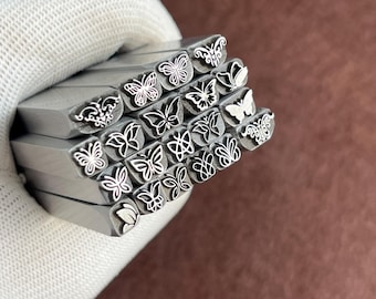 Könnte beinhalten: Eine Sammlung von Metallstempeln mit Schmetterlingsmotiven. Jeder Stempel zeigt ein anderes Schmetterlings- oder Blumenmuster. Die Stempel werden in einem weißen Handschuh vor braunem Hintergrund gehalten.