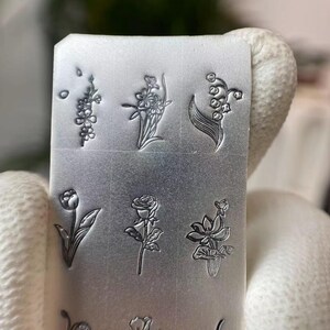 Pu&ograve; includere: Una piastra di metallo argentata con nove diversi disegni di fiori incisi su di essa. I disegni includono mughetto, una rosa, un tulipano, un iris e altri motivi floreali.